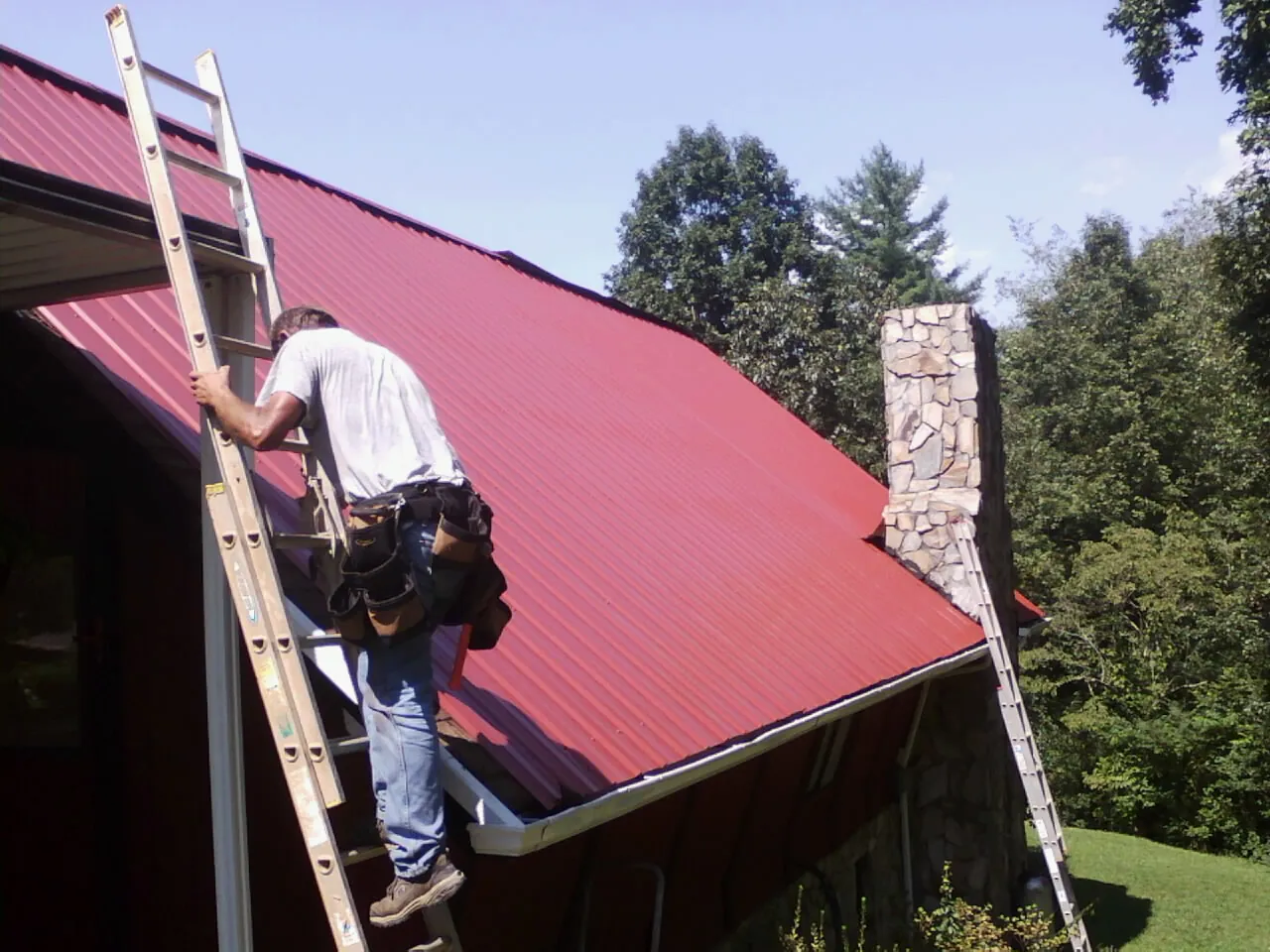 Roofer on a ladder beside a red metal roof and stone chimney — Soffit & Fascia Repair by Roofing Brothers in North Babylon